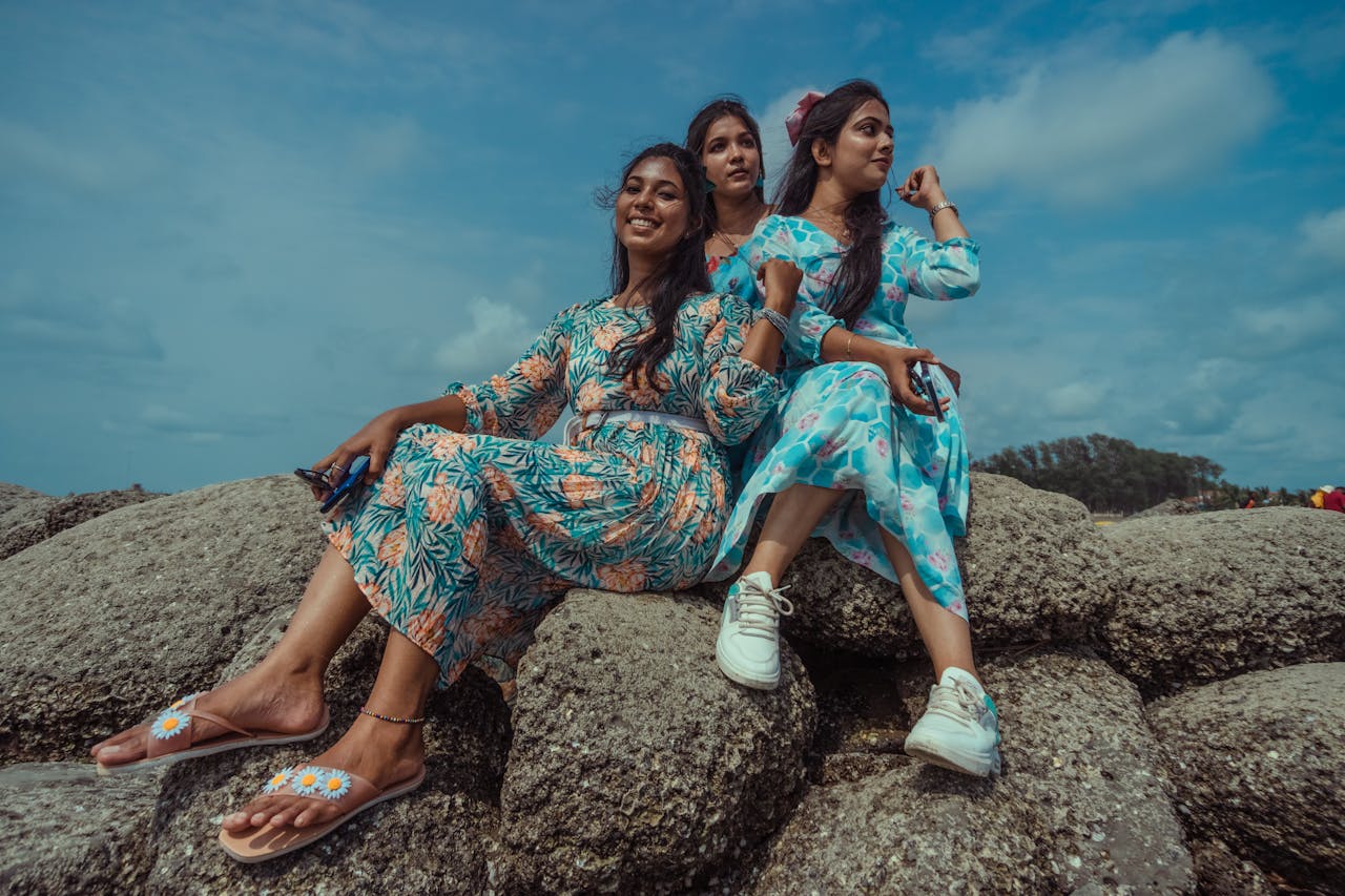 Home Three women enjoy a sunny day sitting on rocks by the sea, wearing colorful floral dresses.