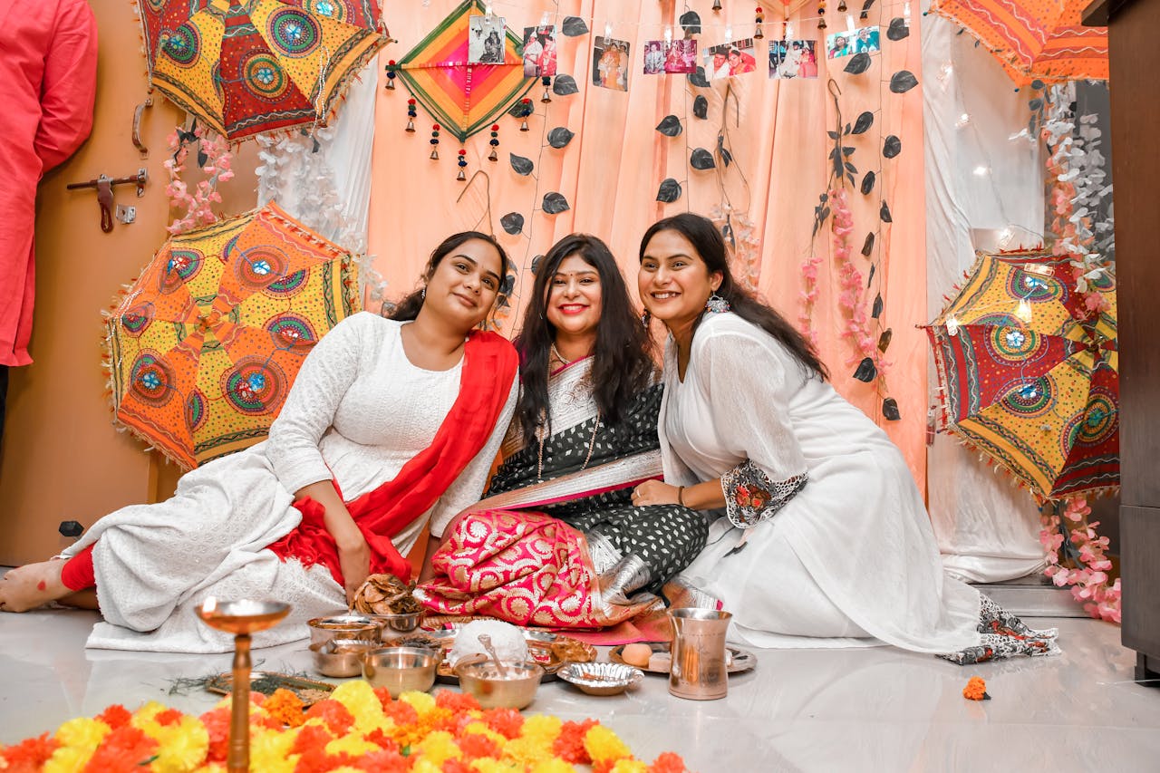 Three women in traditional sarees enjoying a festive gathering indoors with vibrant decor.