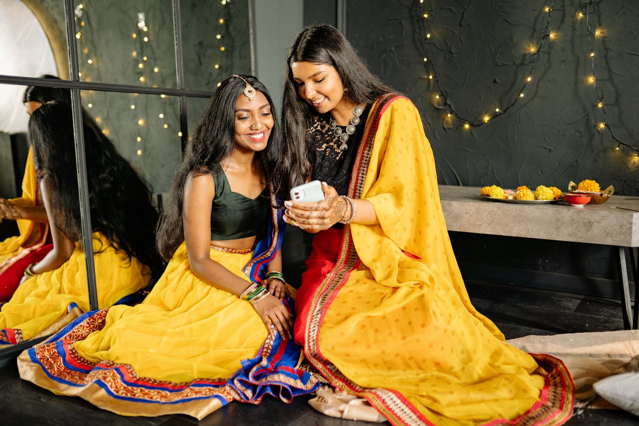 Smiling Indian women in traditional attire enjoying a festive moment with string lights and floral decor.